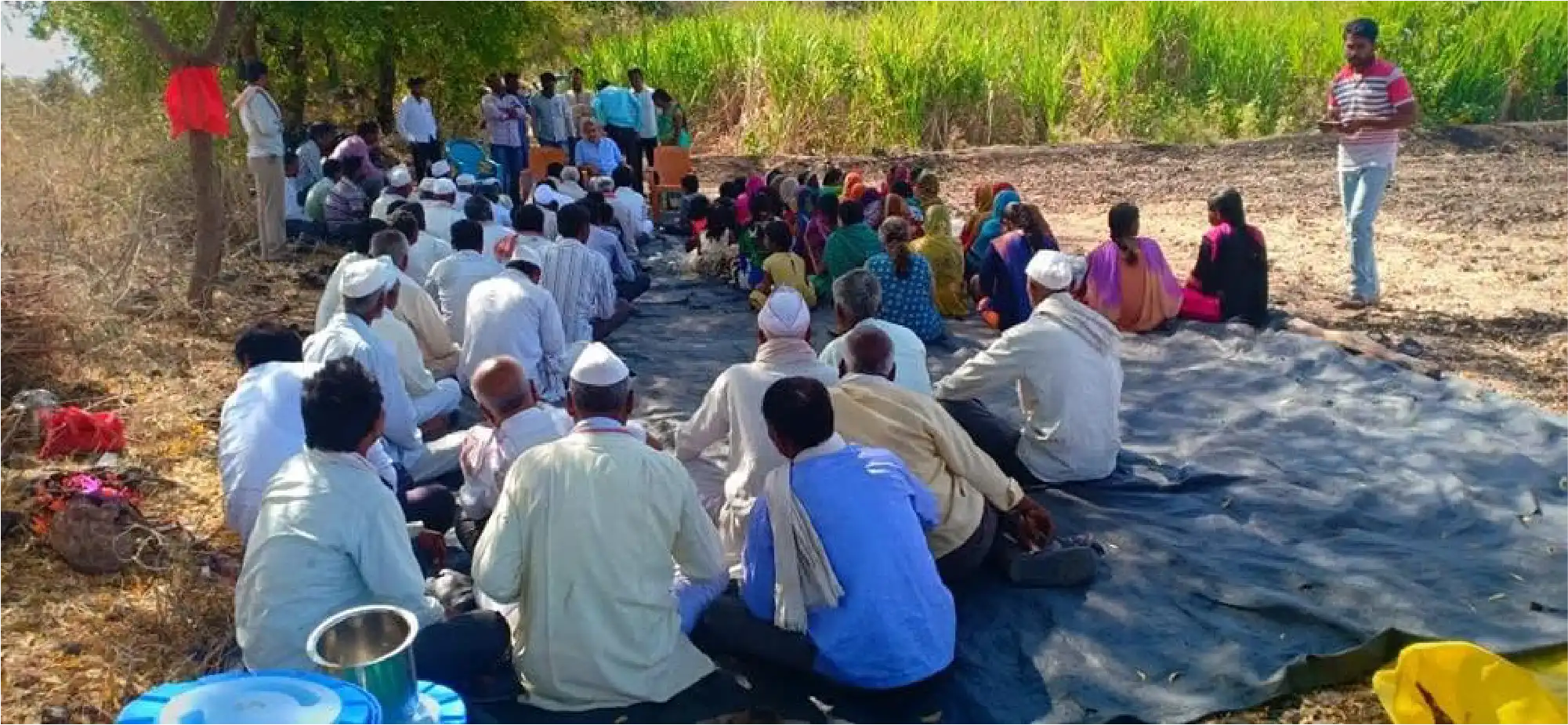 Farmers and village families seated together at an open-air community meeting beside a field